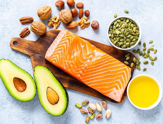 Lipid-rich foods arranged on a board: salmon fillet, avocado halves, walnuts, pecans, hazelnuts, pumpkin seeds, pistachios and a bowl of oil